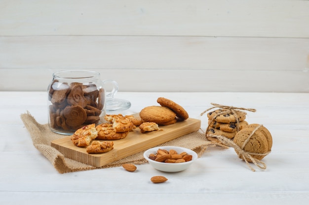 Some brown cookies with almonds in a bowl,cookies on a cutting board and a piece of sack in a glass jar on white surface