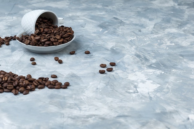 Some coffee beans in cup and plate on grey plaster background, high angle view.