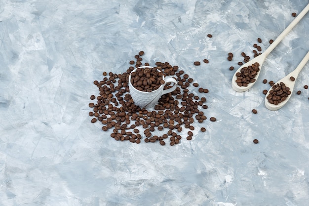 Some coffee beans in white cup and wooden spoons on grey plaster background, high angle view.