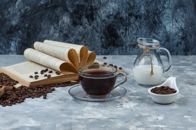Some coffee with coffee beans, book, milk in a cup on grunge and plaster background, side view.