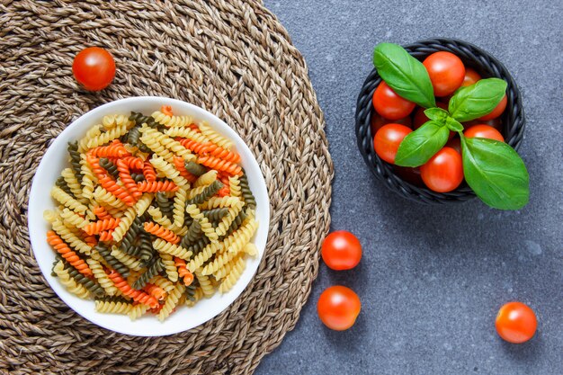 Some colorful macaroni pasta with tomatoes and leaves in a bowl on trivet and gray surface, top view.