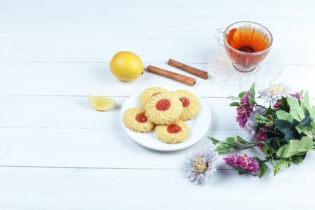 Some cookies, cup of tea with cinnamon, lemon, flowers on white wooden board background, high angle view.