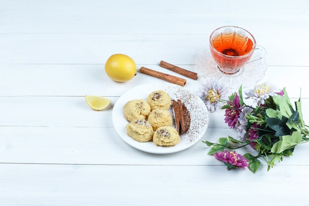 Some cookies, cup of tea with cinnamon, lemon, flowers on white wooden board background, high angle view.