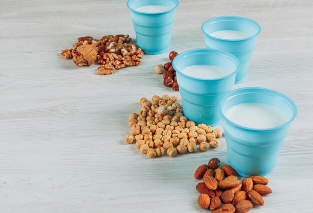 Some cups of milk with hazelnuts, almonds and several nuts on white wooden background, high angle view.