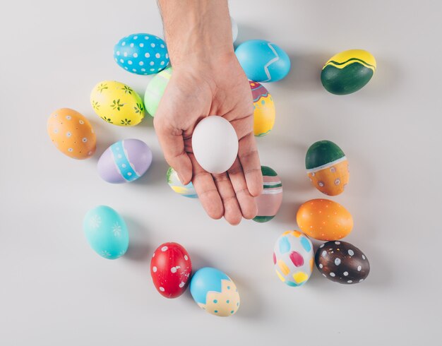 Some easter eggs with man holding one white egg on white background, top view.