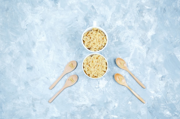 Alcune farfalle di pasta in ciotole e cucchiai di legno su sfondo di gesso, vista dall'alto.