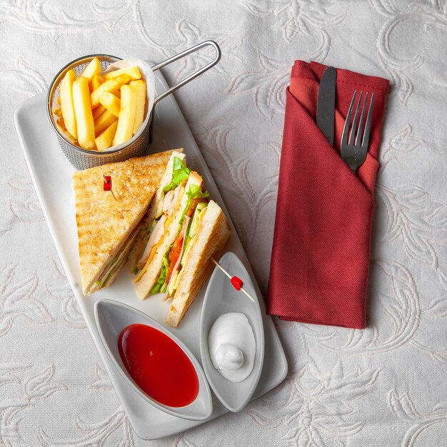 Some fast food with sandwich, french fries, fork and knife on white textured background, top view.