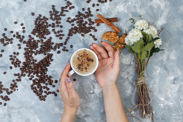 Некоторые женские руки держат чашку кофе с кофейными зернами, палочками корицы, цветами, печеньем на сером гипсовом фоне, плоской планировкой.
