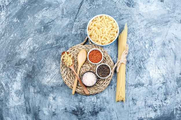 Free photo some fusilli pasta with spaghetti, wooden spoons, spices in a bowl on grey plaster and wicker placemat background, top view.