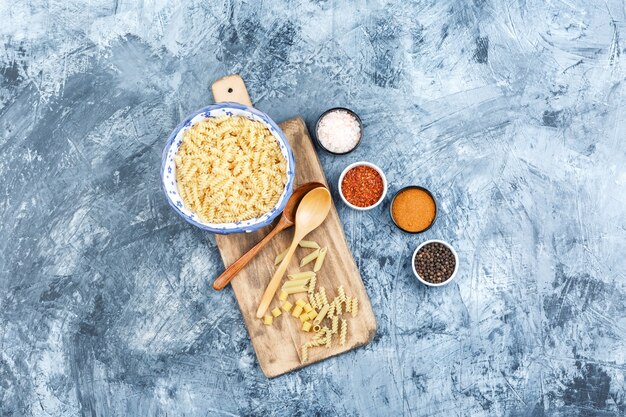 Some fusilli pasta with spices, wooden spoons in a bowl on grey plaster and cutting board background, top view.