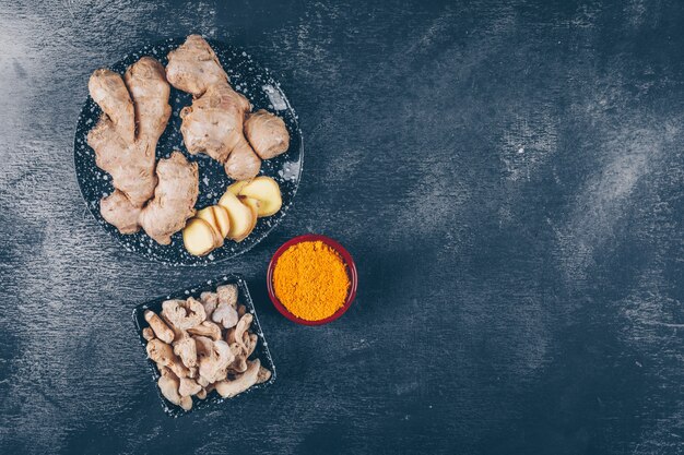 Some ginger with ginger slices and powder in bowls and plate on dark textured background, top view. space for text