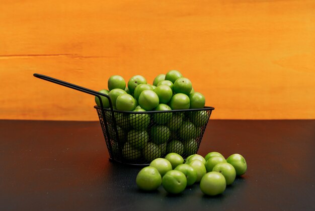 Some green cherry plum with several green cherry plum around it in a black iron bowl on orange background, high angle view.