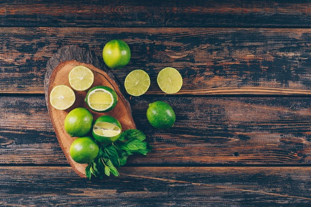 Some green lemons with slices and leaves on wood slice and dark wooden background, flat lay. space for text