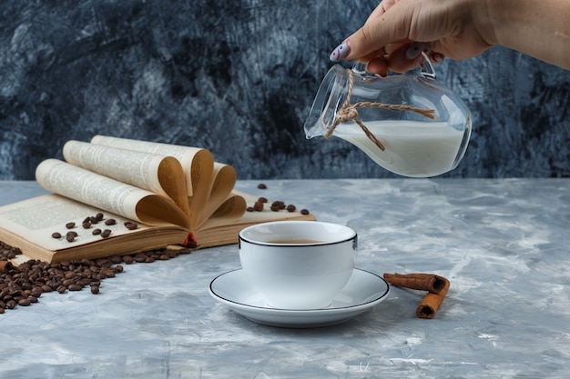 Some hand pouring milk into a cup of coffee with coffee beans, cinnamon sticks, book on grunge and plaster background, side view.