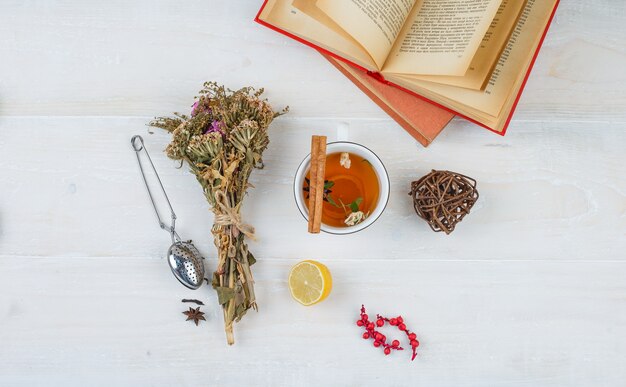 Some herbal tea and flowers with books,lemon,tea strainer and spices on white surface