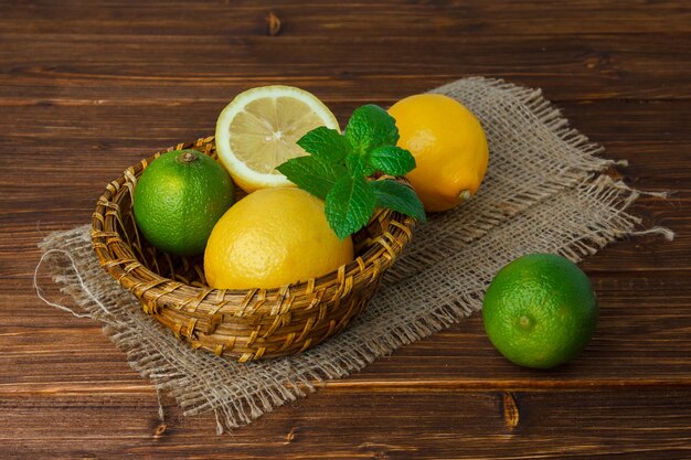 Some lemon and leaves with half of lemon on piece of sack in a basket on wooden surface, high angle view. space for text