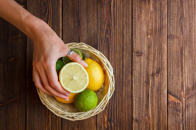 Some lemons with hand holding half of lemon in a basket on wooden background, top view.