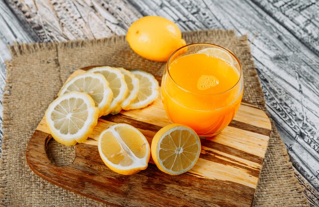 Some lemons with slices and juice on cutting board, cloth and wooden background, high angle view.