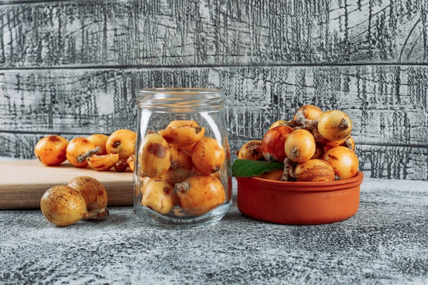Some loquats in a jar, bowl and cutting board on gray textured background, side view.