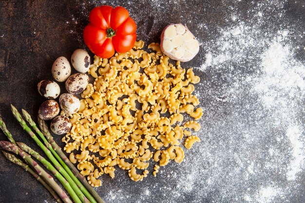 Some macaroni with eggs, tomato, asparagus and garlic on dark textured background, top view.