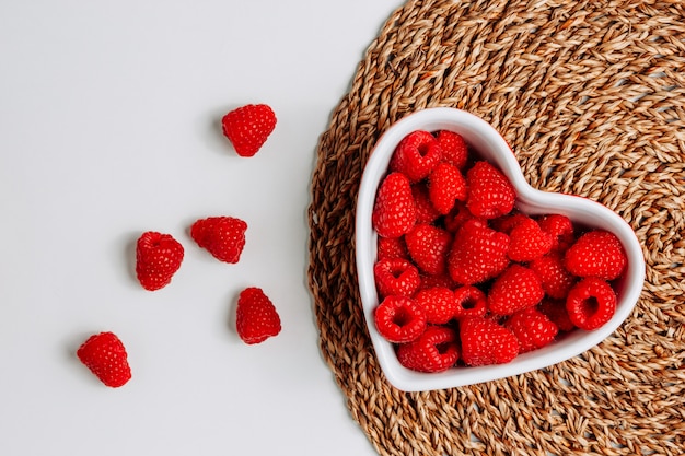 Some raspberries in a heart shaped bowl on trivet and white background, top view.