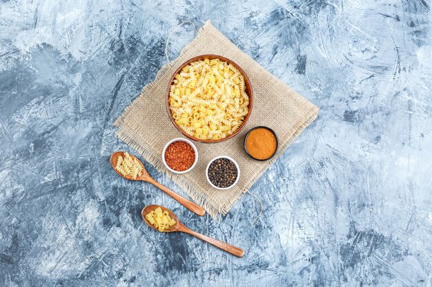 Some raw pasta with spices in bowl and wooden spoons on plaster and piece of sack background, top view.