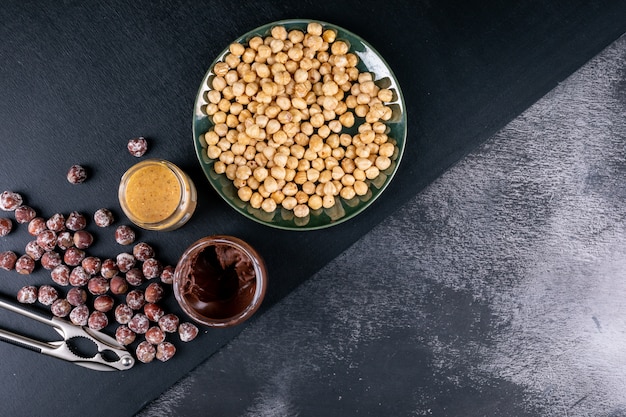 Some of shelled and cleaned hazelnuts with cocoa spread and nutcracker in a glassy plate on dark stone table, top view.