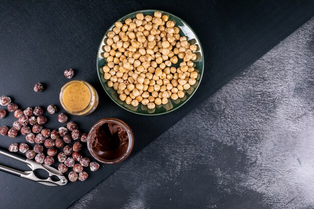 Some of shelled and cleaned hazelnuts with cocoa spread and nutcracker in a glassy plate on dark stone table, top view.