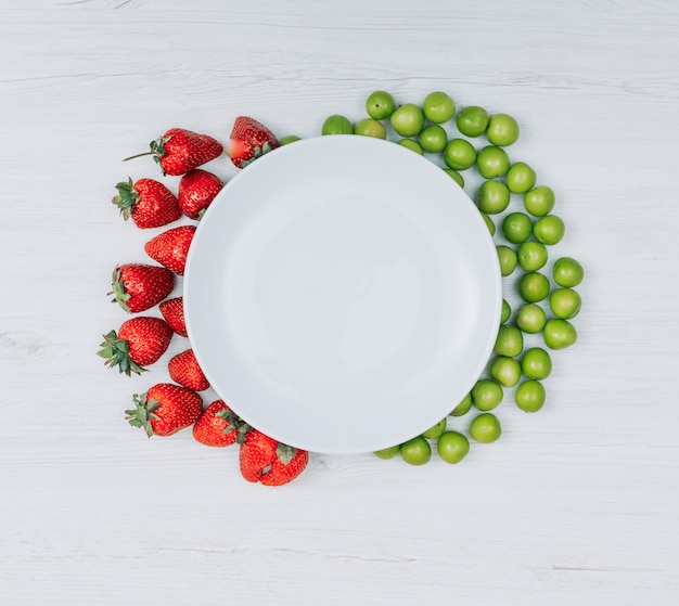 Some strawberries and green cherry plums with empty plate on white wooden background, flat lay. copy space for text