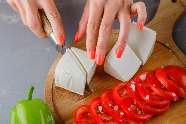 Some woman slicing cheese with sliced tomatoes, on a cutting board on gray surface