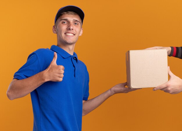 Кто-то дает картонную коробку улыбающемуся молодому светловолосому мальчику-доставщику, который поднимает палец вверх изолированно на оранжевой стене с копией пространства