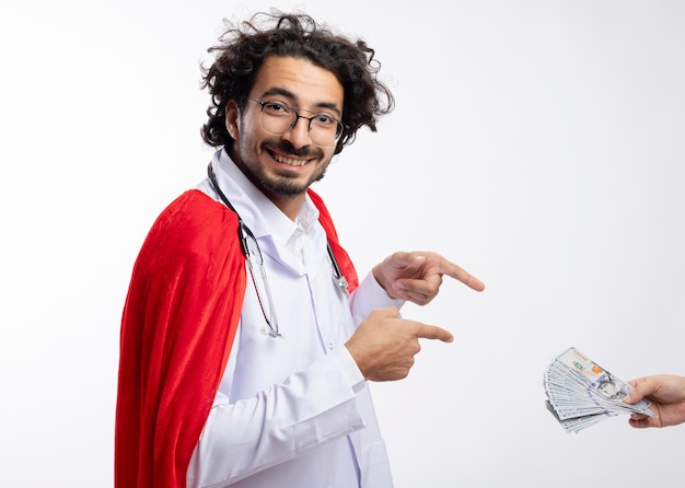 Someone hands money to smiling young caucasian man in optical glasses wearing doctor uniform with red cloak and with stethoscope around neck pointing at money isolated on white wall