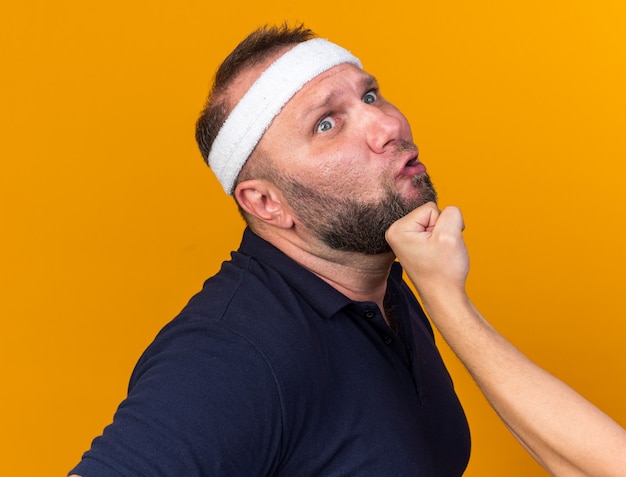 Someone punching in chin adult slavic sporty man wearing headband and wristbands isolated on orange wall with copy space