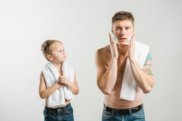 Free photo son looking at his father applying the shaving foam over his face against grey backdrop