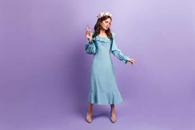 Sophisticated European lady enjoying glass of champagne on purple wall. Full-length photo of dark-haired model in blue dress.