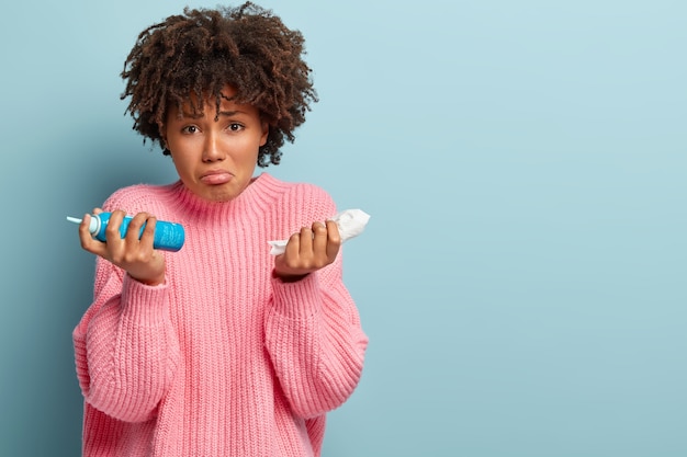 Sorrowful woman suffers from seasonal allergy, holds handkerchief and nasal aerosol, has Afro hairstyle, wears oversized pink jumper, models over blue wall with blank space for your text