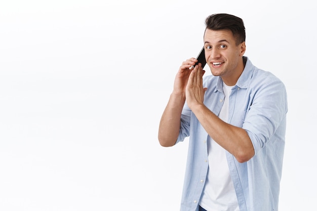 Sorry cant talk right now. Portrait of smiling handsome man busy talking on phone, hold mobile phone and cover dynamic to say something to you, having conversation, white wall