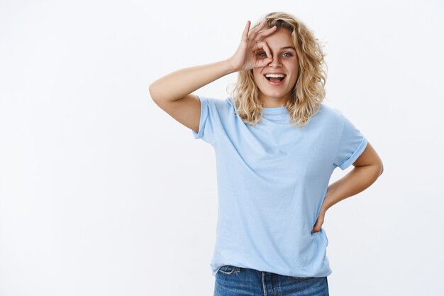 Sounds awesome, okay. Portrait of happy and playful grinning cute blond girl with blue eyes in t-shirt holding hand on waist and showing ok gesture on eye, smiling at perfect idea