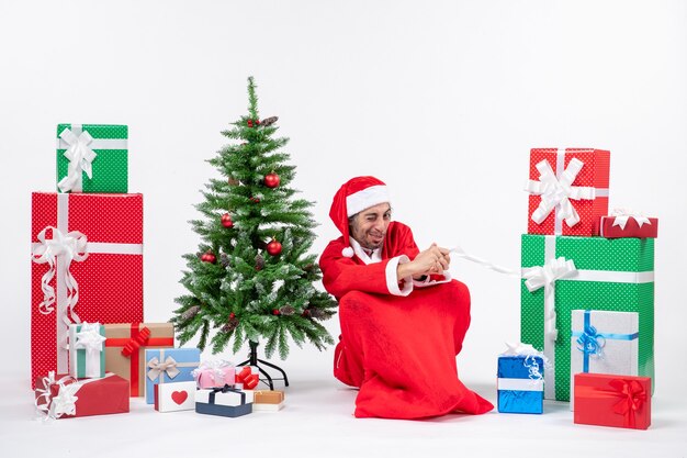 Sour face young man dressed as Santa claus with gifts and decorated Christmas tree sitting on the ground on white background
