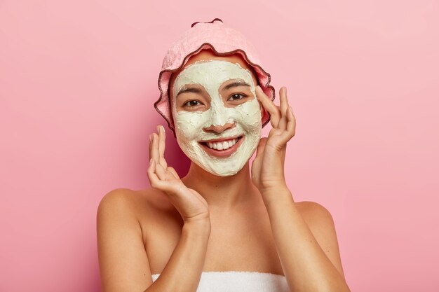 Spa facial mask application. Happy delighted young woman with Asian appearance, improves skin condition with clay mud, applies cosmetic product on face, takes care of her complexion and body