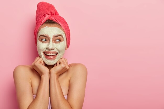 Spa girl smears muddly texture across face, has glad look, keeps hands under chin, looks away with smile, hydrates and calms skin, wears red towel on head, excess oil production, stands naked indoor