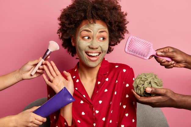 Free photo spa at home. glad dark skinned young woman raises palm, dressed in red pajama, looks gladfully aside, applies clay mask