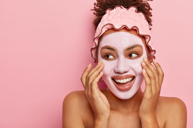 Spa and skin care concept. Pleased Afro American woman applies nourishing clay mask on face, has glad expression, looks on left side, touches cheeks, fights with problem of dry skin, has topless body