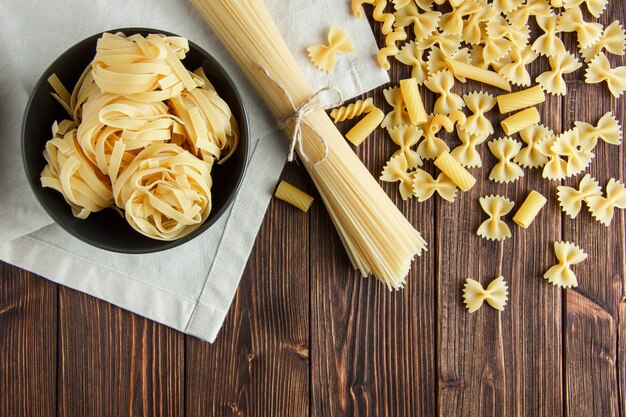 Spaghetti with assorted raw pasta on wooden and kitchen towel background, flat lay.