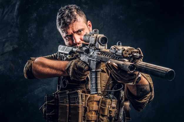 Foto gratuita soldato delle forze speciali che indossa un'armatura che tiene un fucile d'assalto e mira al nemico. foto dello studio contro una parete strutturata scura