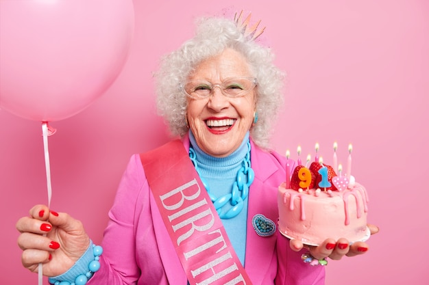 Special occasion age and festivity concept. Happy smiling wrinkled senior woman holds festive strawberry cake inflated balloon prepares for party or birthday celebration expresses good emotions