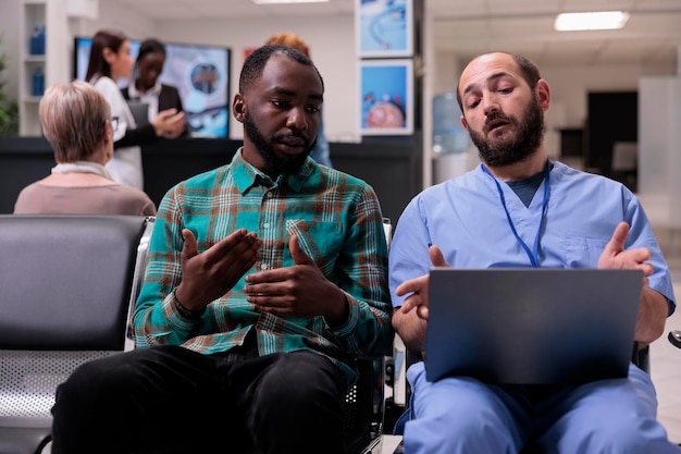 Специалист консультирует афроамериканца в зоне ожидания, используя ноутбук, чтобы объяснить диагноз заболевания и восстановительное лечение в холле стойки регистрации. Люди говорят о здравоохранении в учреждении.