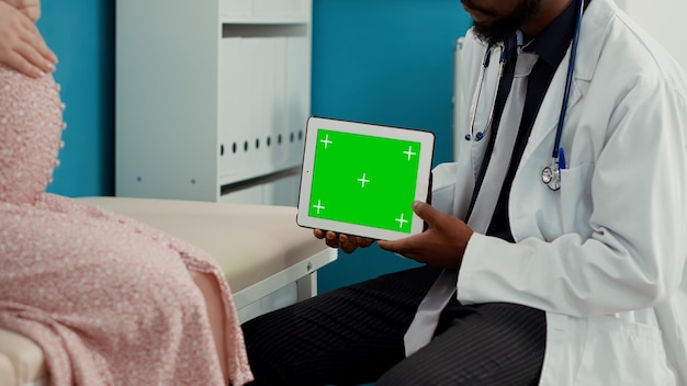 Specialist holding digital tablet with horizontal greenscreen in medical cabinet. Medic and pregnant woman looking at blank isolated chroma key with copyspace mockup template. Close up.