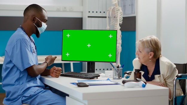 Specialist and old woman using greenscreen monitor in cabinet, patient with physical disability. analyzing blank mockup template with isolated copyspace and chromakey display. tripod shot.