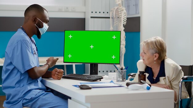 Специалист и пожилая женщина, использующая монитор с зеленым экраном в кабинете, пациент с ограниченными физическими возможностями. Анализ пустого шаблона макета с изолированным копирайтом и отображением хромакея. Съемка со штатива.
